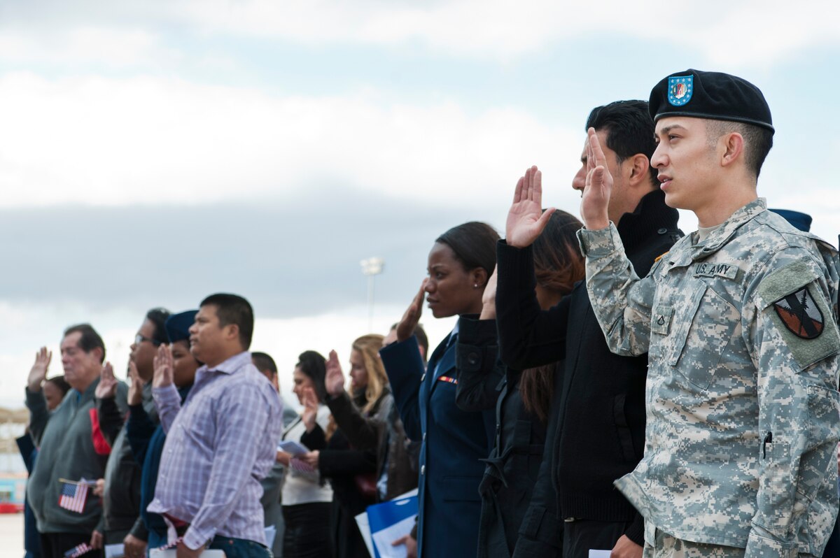 A man in an Army uniform raises his right hand together with a large group of people.