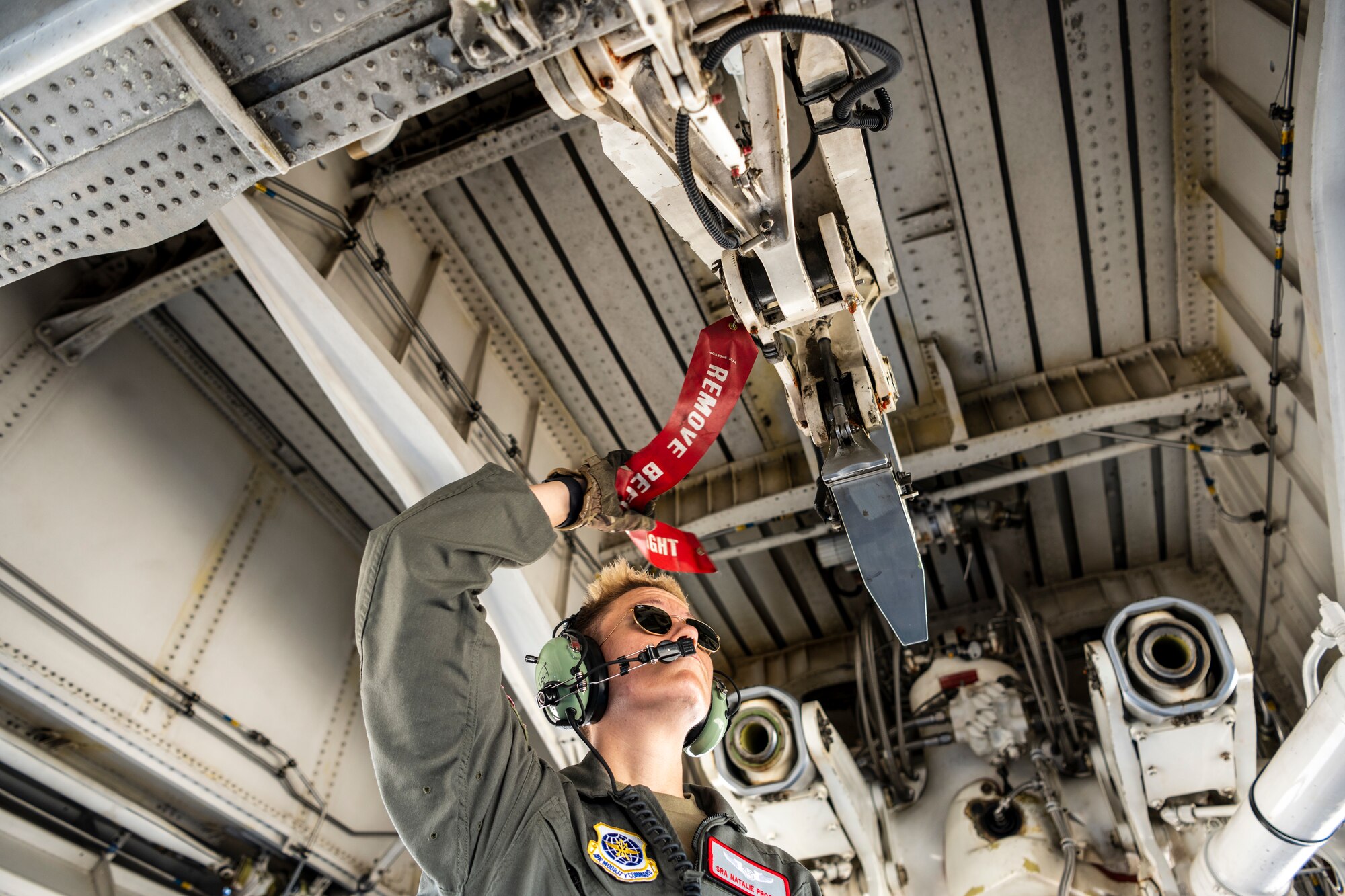 A woman in a flight suit places a red flag that says remove before flight below a large military airplane.