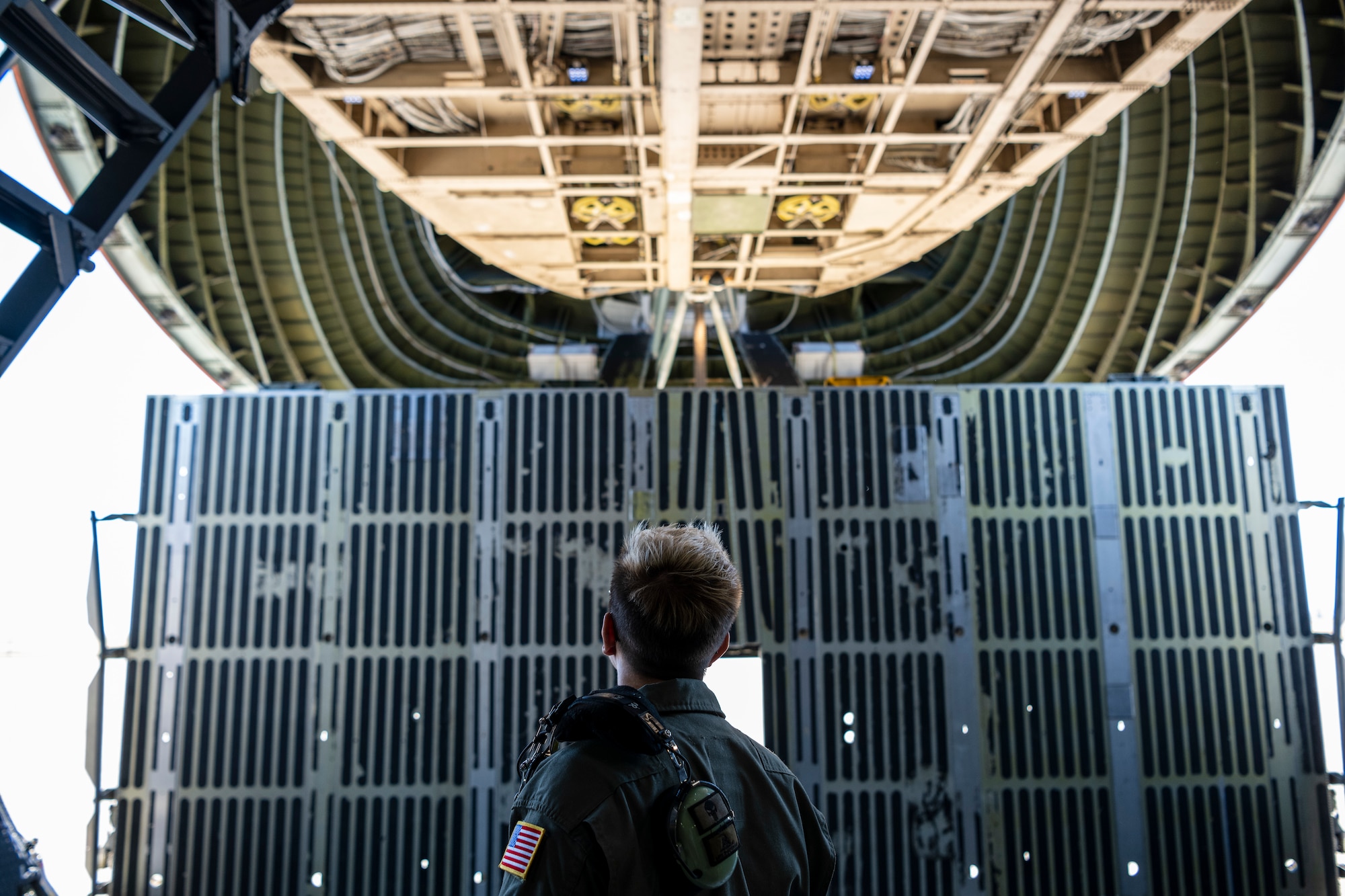 A woman in a flight suit watches a large door open on a large military airplane.