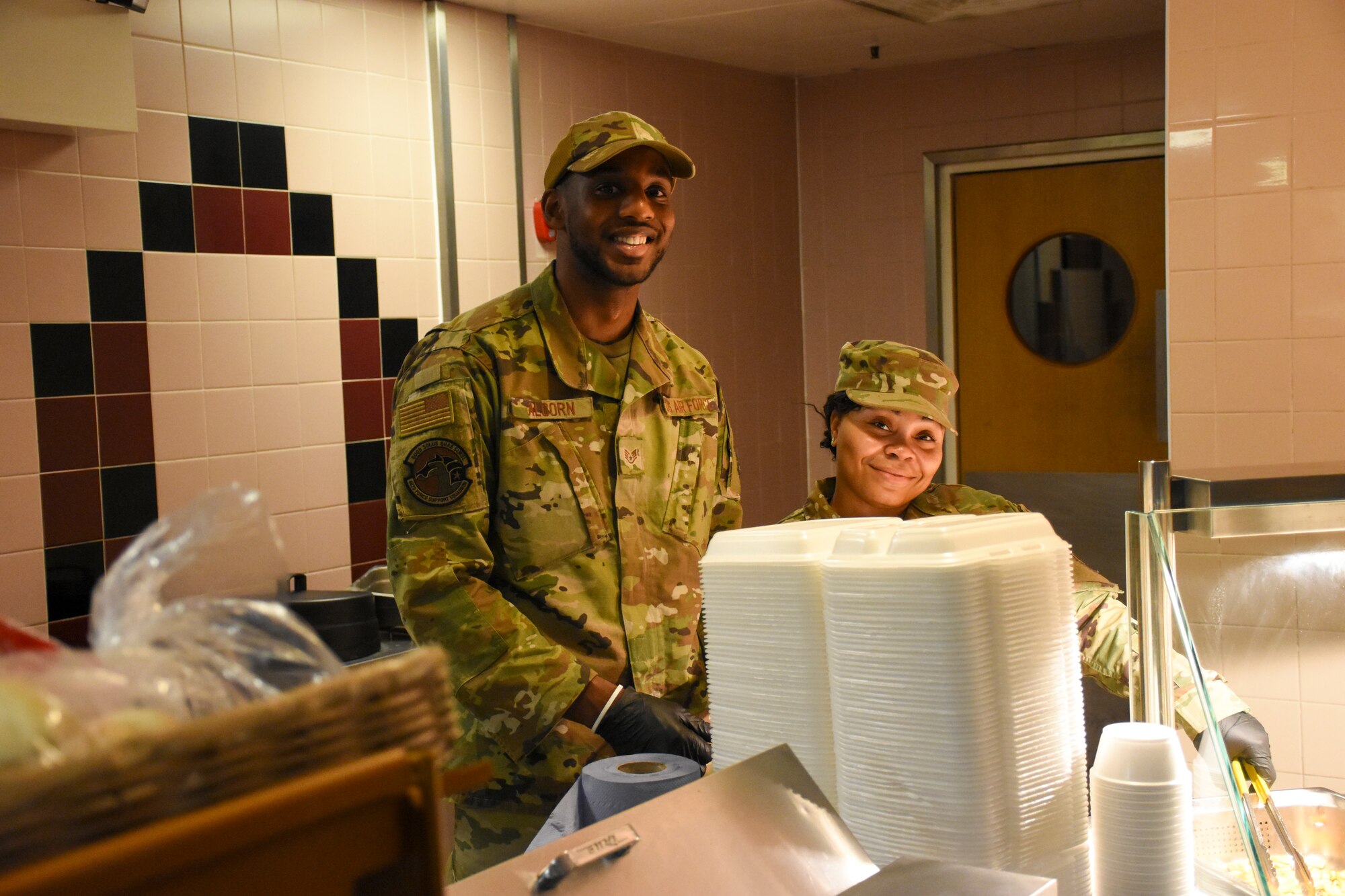 A very short woman and a very tall man stand behind a food service counter. The woman is hidden almost entirely behind a stack of Styrofoam serving trays.