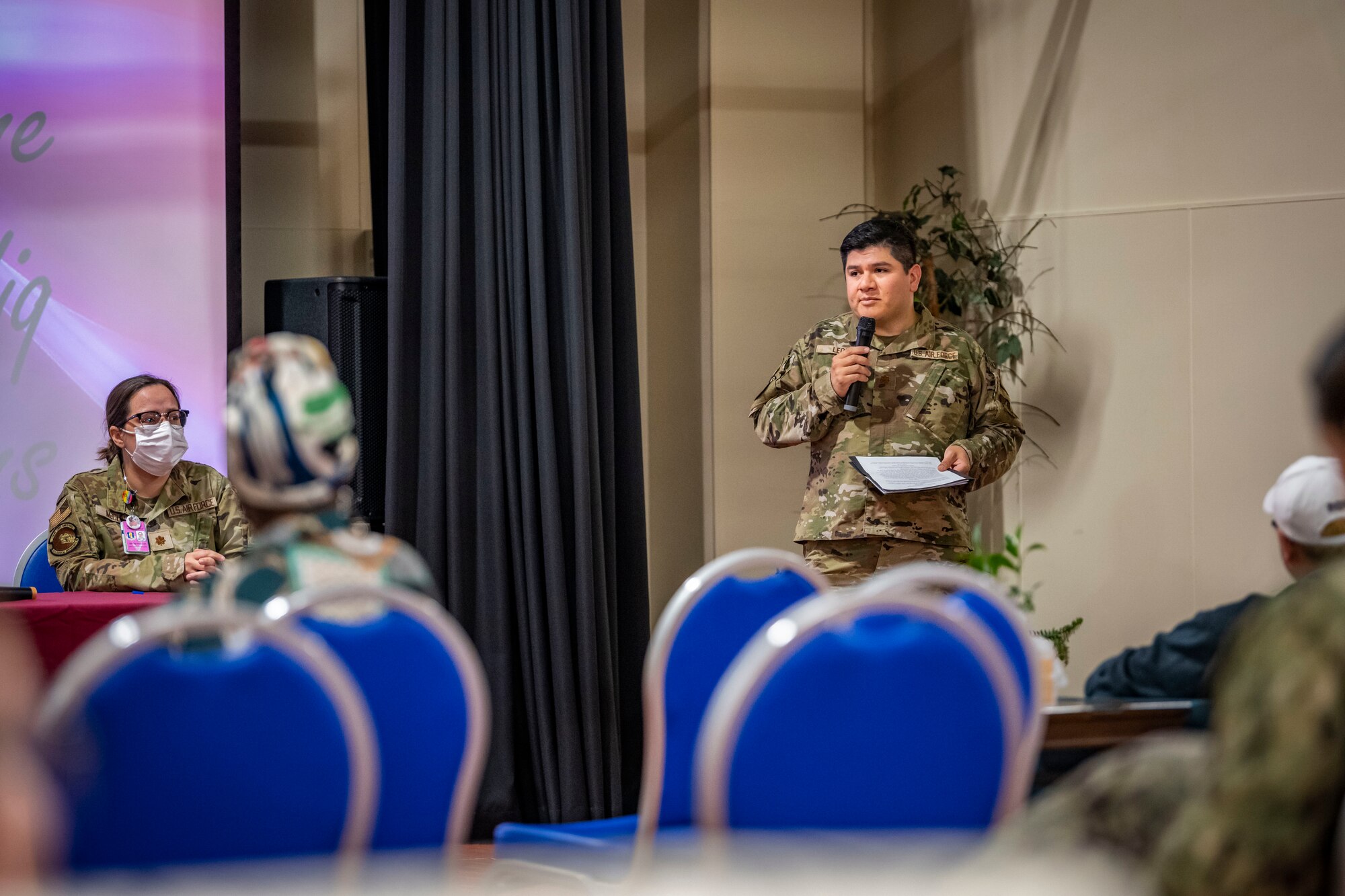 Military member in uniform talks into a microphone towards a crowd of people.