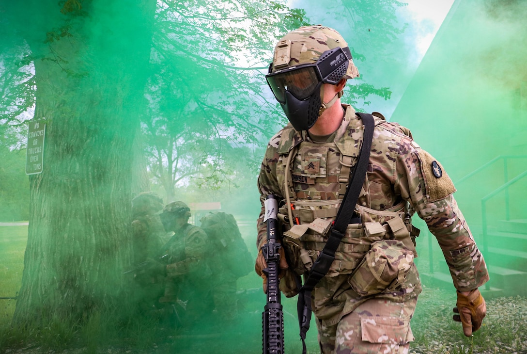 A soldier walks through a wooded area surrounded by green smoke.