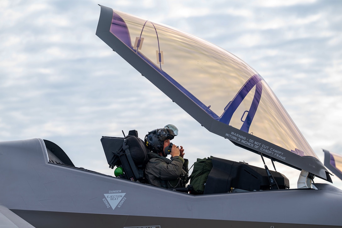 A pilot sits in the cockpit of a jet with the canopy open while holding an oxygen mask to his mouth.