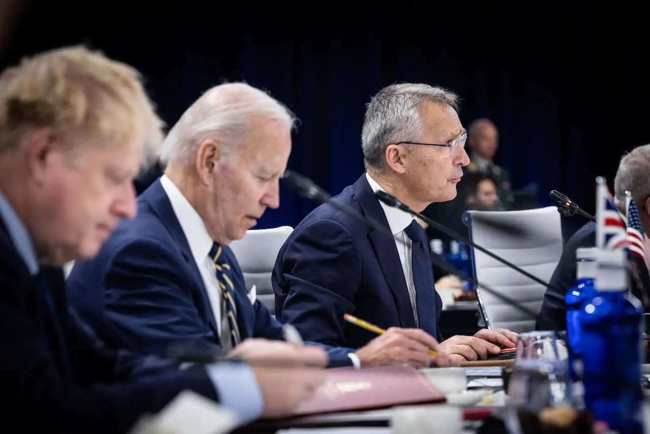 A man speaks into a microphone; two other men sit to his right.