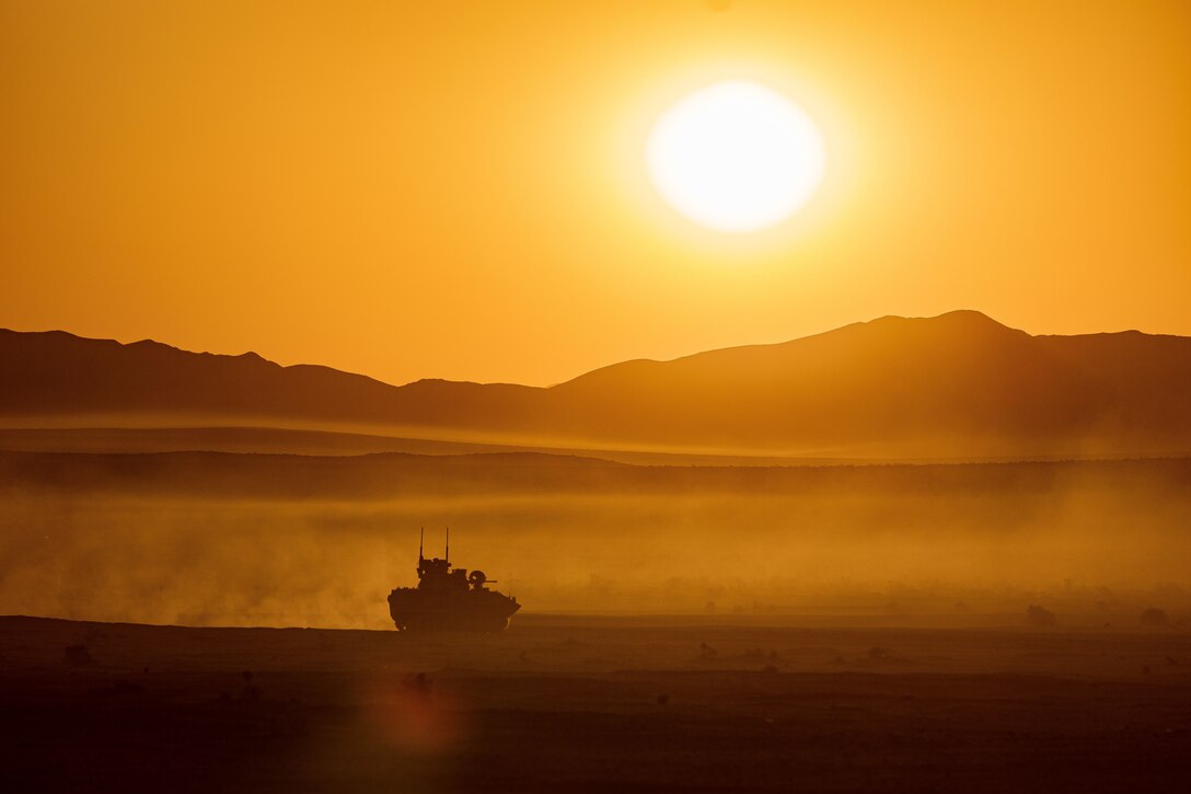 An armored vehicle drives in a desert-like area under a sunlit sky.