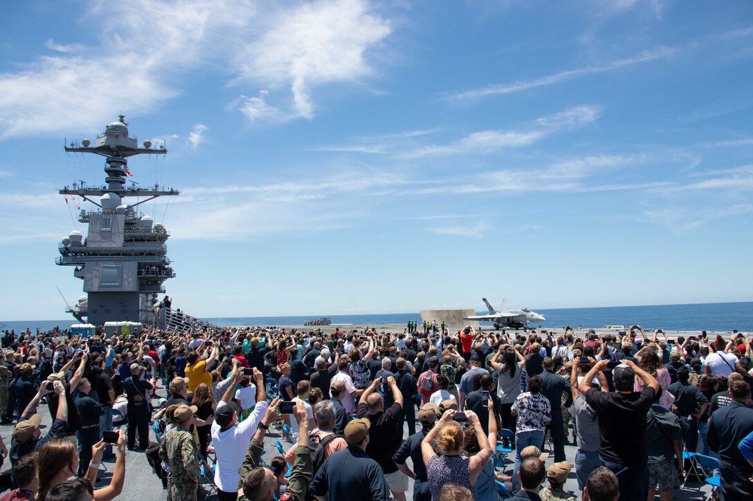 A crowd of people gather and wave form the deck of an aircraft carrier.