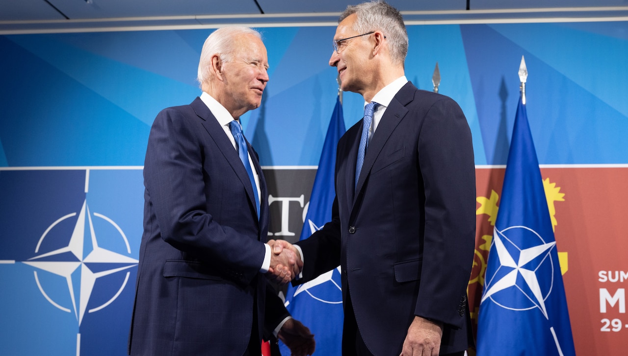 Two men sit in chairs conversing. There are flags in the background.