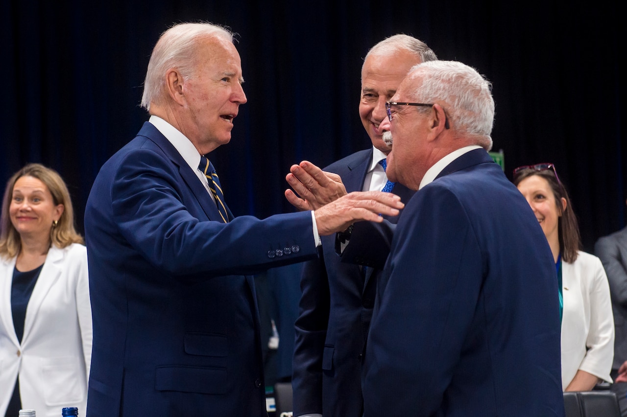 Three men in business attire talk to each other while several other look on in the background.