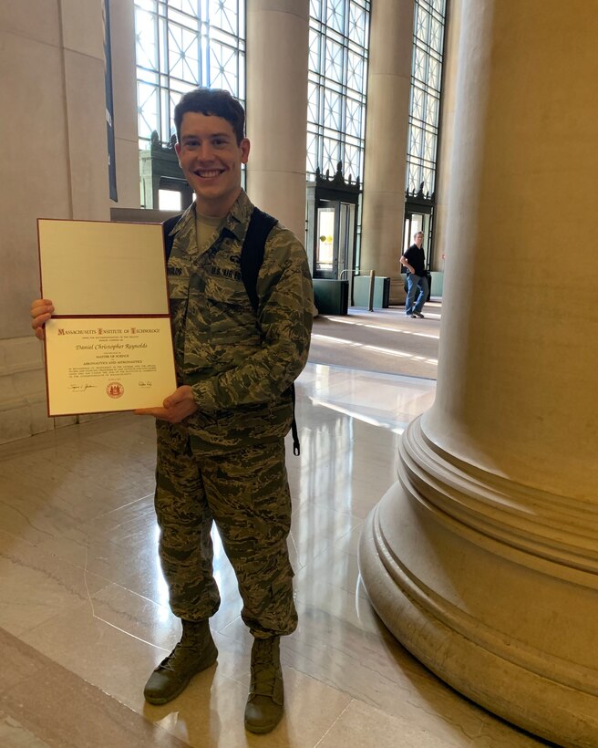An airman displays a diploma while standing in a large building foyer.