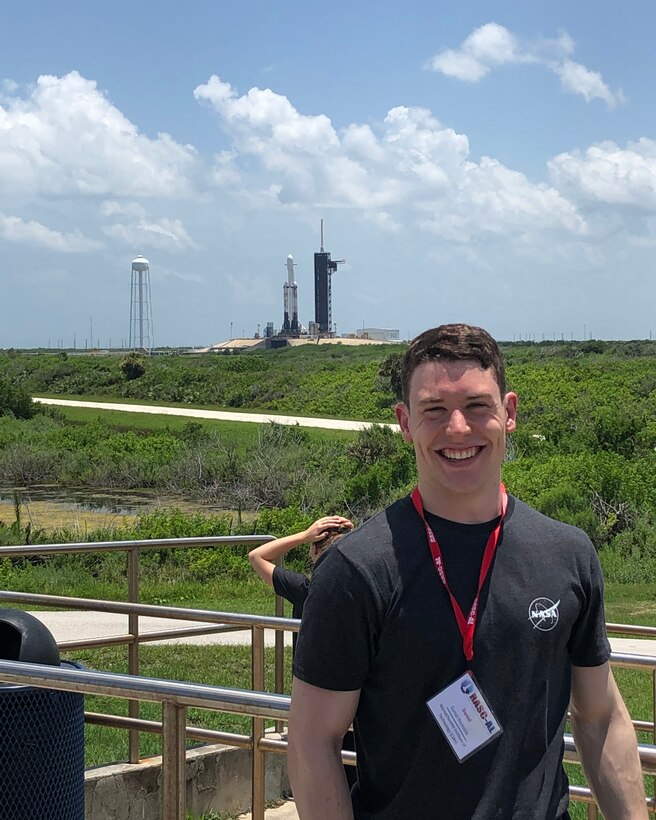 A man poses in front of a rocket on a launch pad in the distance.
