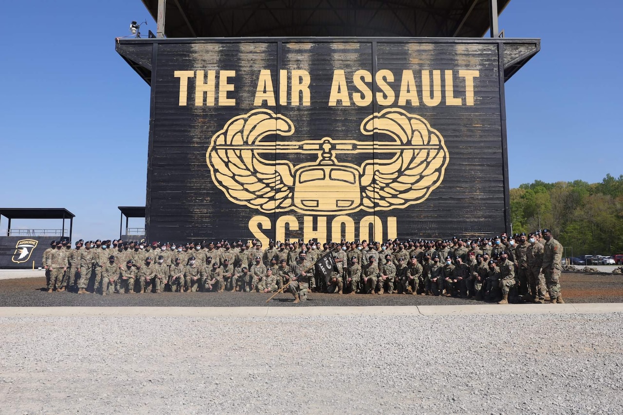 Service members pose for a photo in front of a giant outdoor tower with "The Air Assault School" on it.