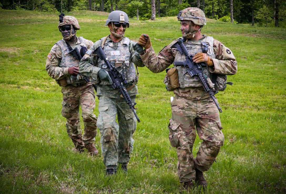 A soldier fist bumps another while walking across a field; a third soldier follows behind.