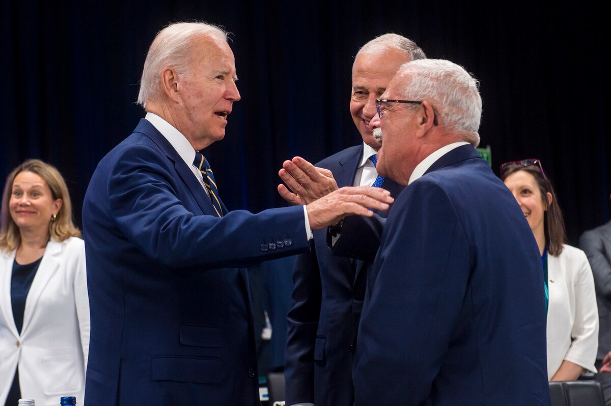 Three men in business attire talk to each other while several other look on in the background.