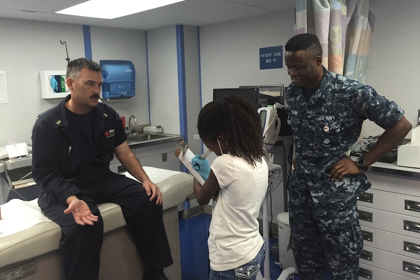 A young girl writes on a clipboard as she pretends to examine a man on a medical table. Another man supervises.