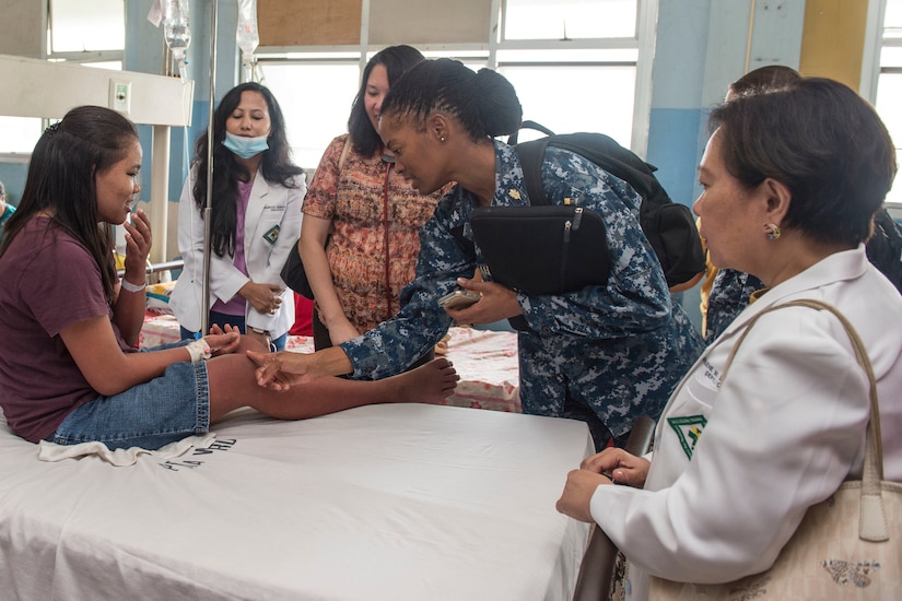 A woman touches the knee of a teen girl sitting on a medical table. Three others watch.