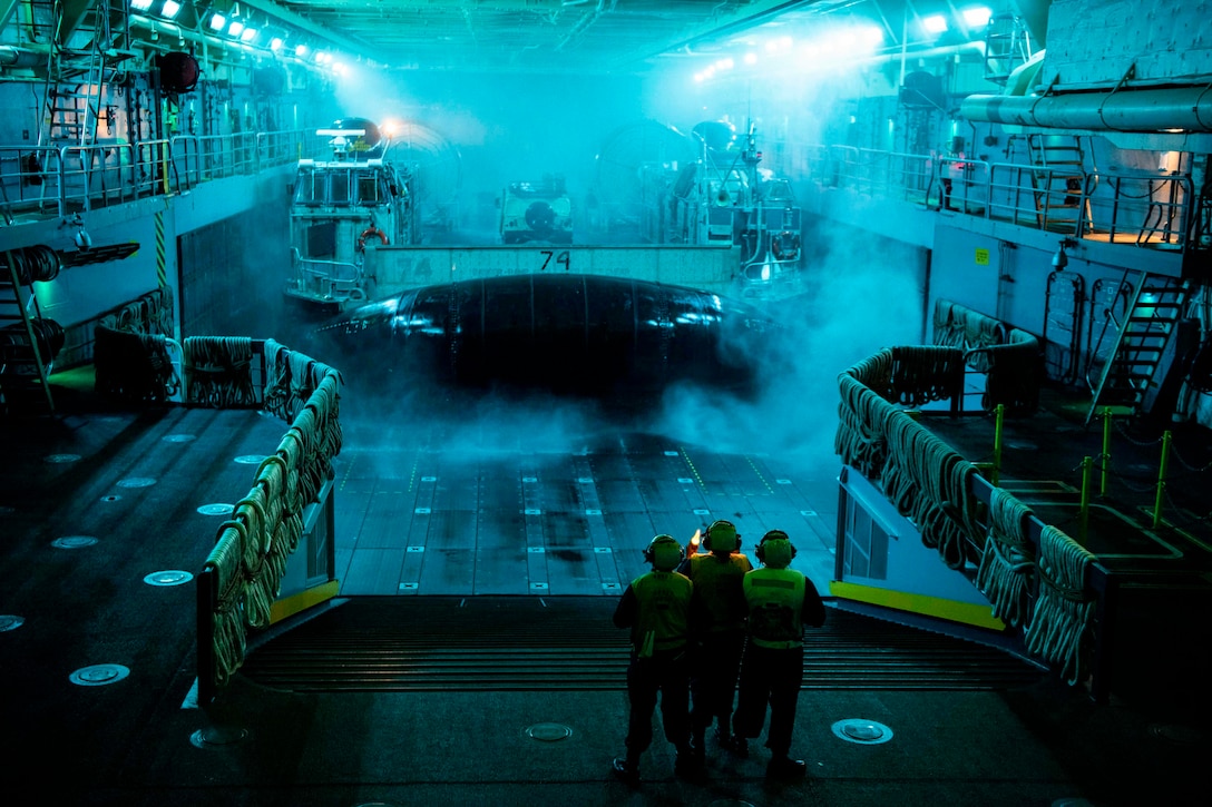 Sailors stand in the well deck of a ship at sea.