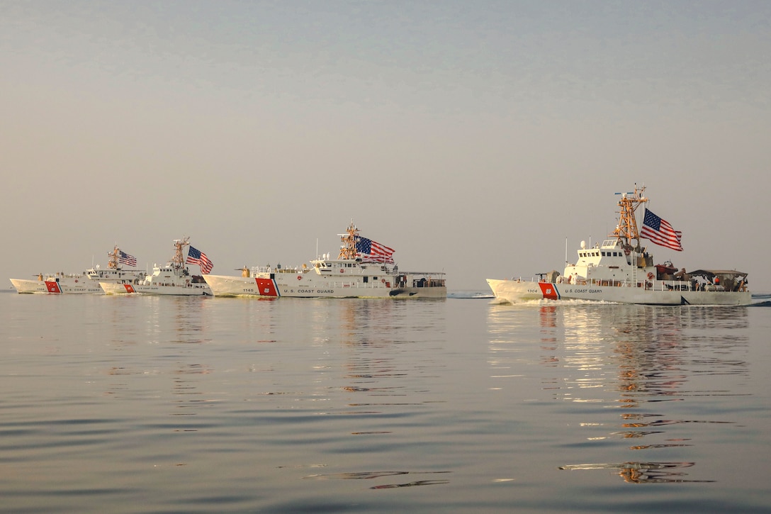 Four ships displaying American flags sail in formation in a body of water.