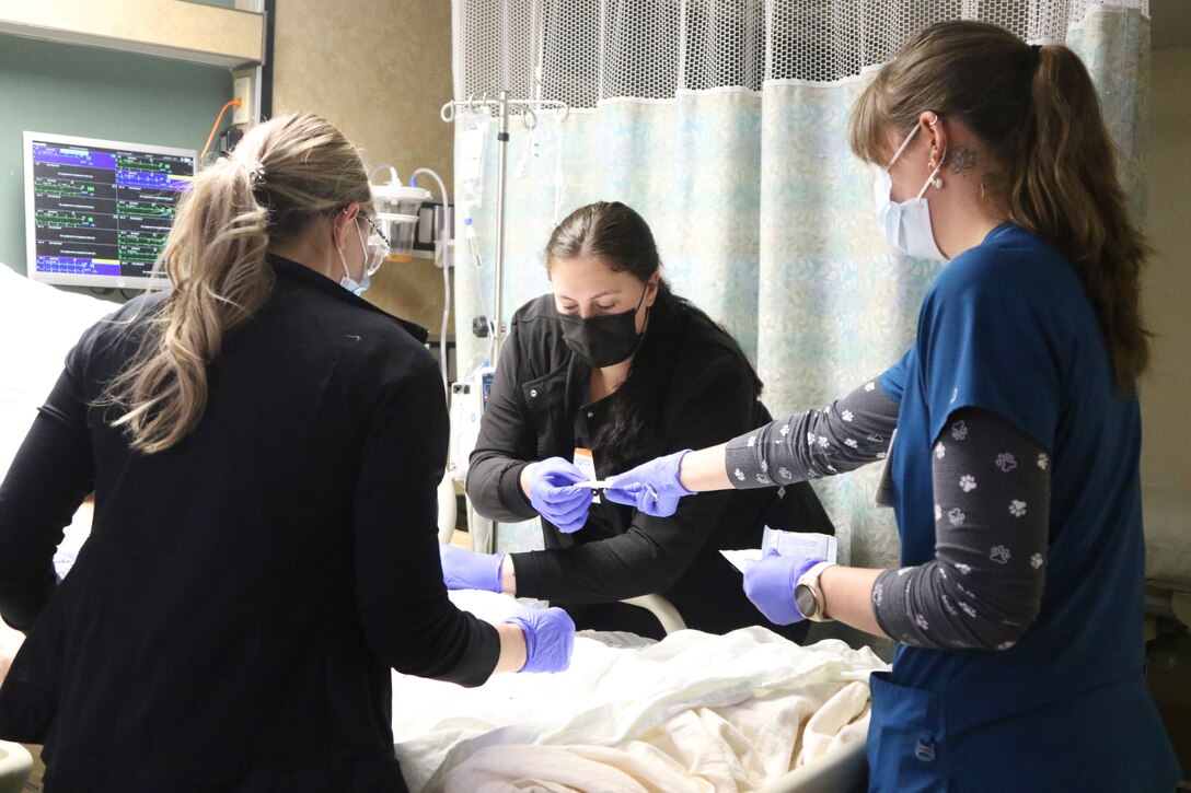 Two Army soldiers and a nursing student, wearing face masks and gloves, work together to fix a patient’s IV.