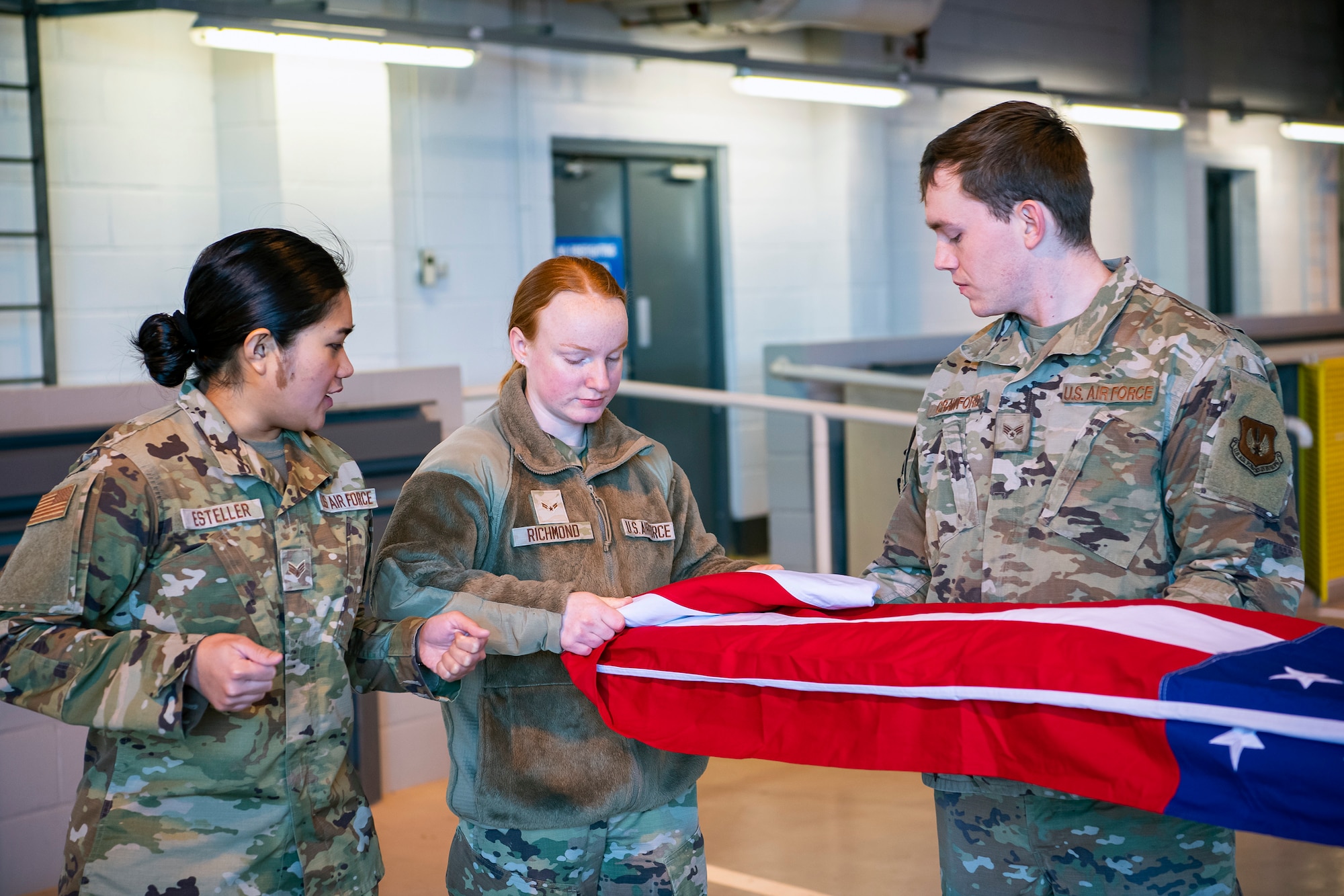 U.S. Air Force Senior Airman Bloom Esteller, center, 423rd Medical Squadron dental assistant, gives instruction on flag folding to Airman 1st Class Alyssa Richmond, 423rd Communications Squadron cyber surety apprentice at RAF Alconbury, England, Feb. 9, 2022. Airmen from the 423 Air Base Group honor guard conduct weekly training sessions to hone their skills and build chemistry in executing details with Airmen from different squadrons. (U.S. Air Force photo by Senior Airman Eugene Oliver)