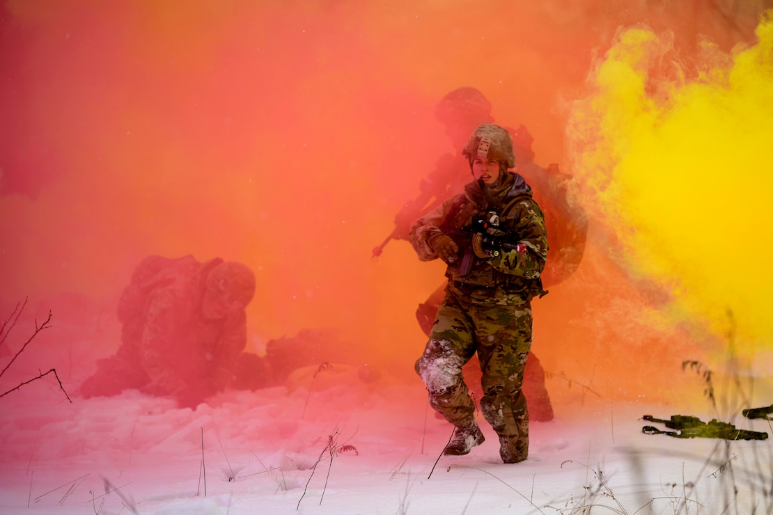 Airmen holding weapons walk through snow and clouds of pink and yellow smoke.