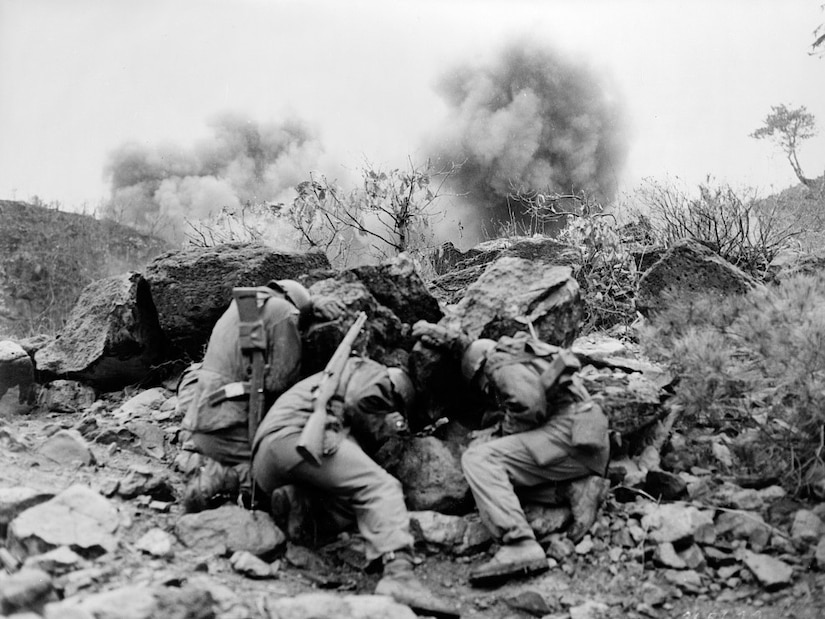 Three men duck for cover behind some boulders as something explodes in the background.