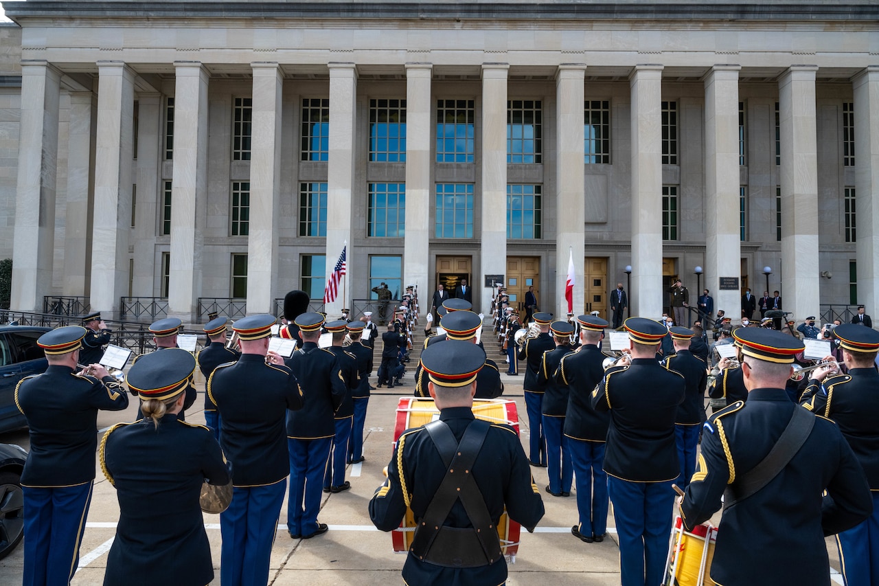 Secretary of Defense Lloyd J. Austin III greets Bahraini Crown Prince Salman bin Hamad Al Khalifa in front of a building as a band performs.