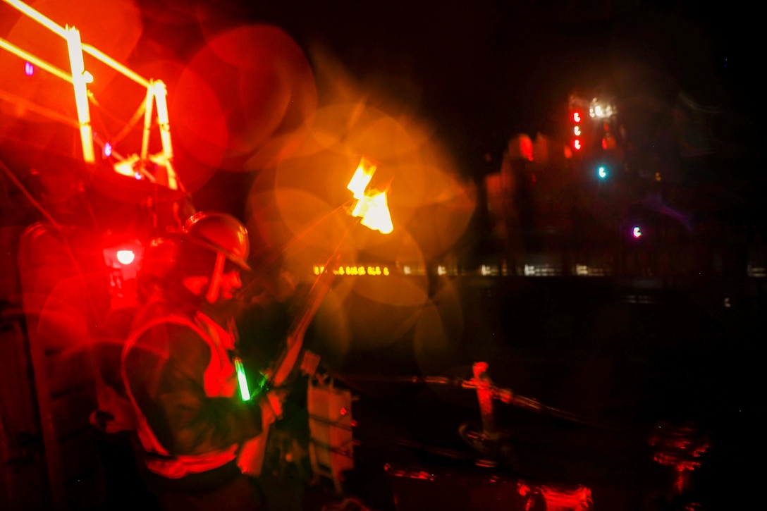 A sailor holds a weapon while standing on a ship as another ship floats nearby illuminated by colorful lights.