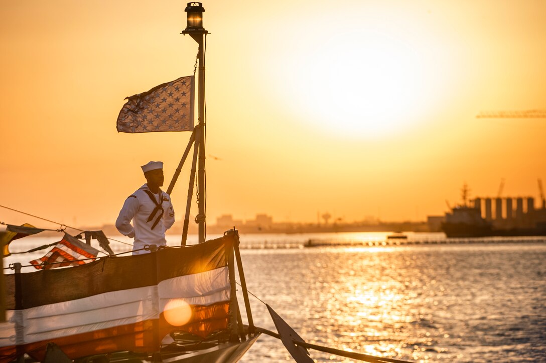 A sailor stands at parade rest aboard a ship at sea under a sunlit sky.