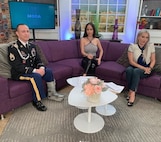 Soldier and 2 news anchors sit on a couch in a news studio.