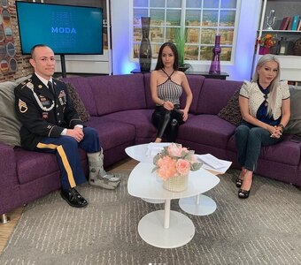 Soldier and 2 news anchors sit on a couch in a news studio.