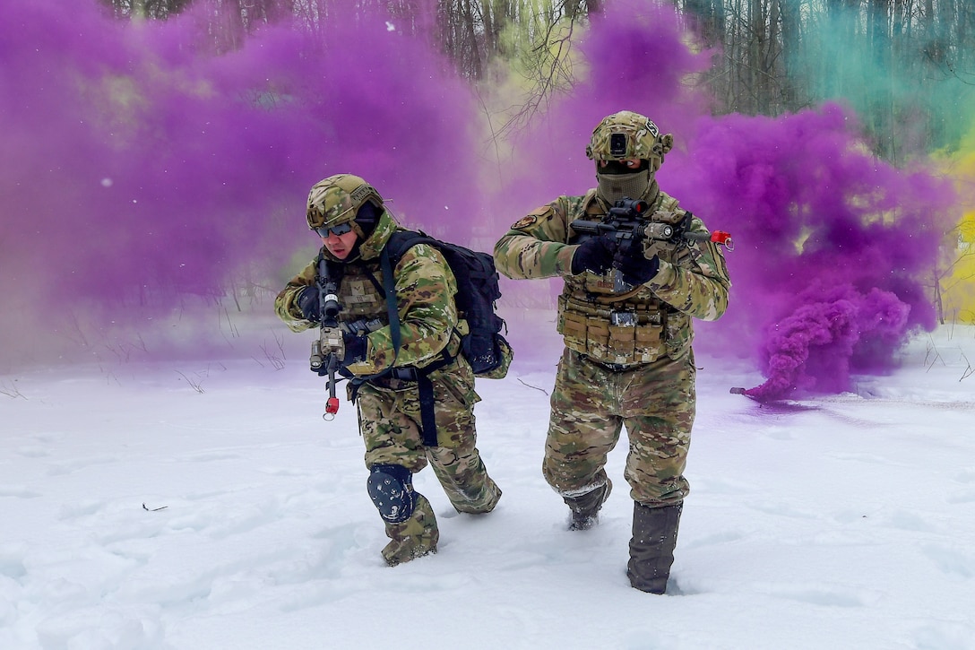 Two airmen run toward the camera in the snow with purple smoke behind them.