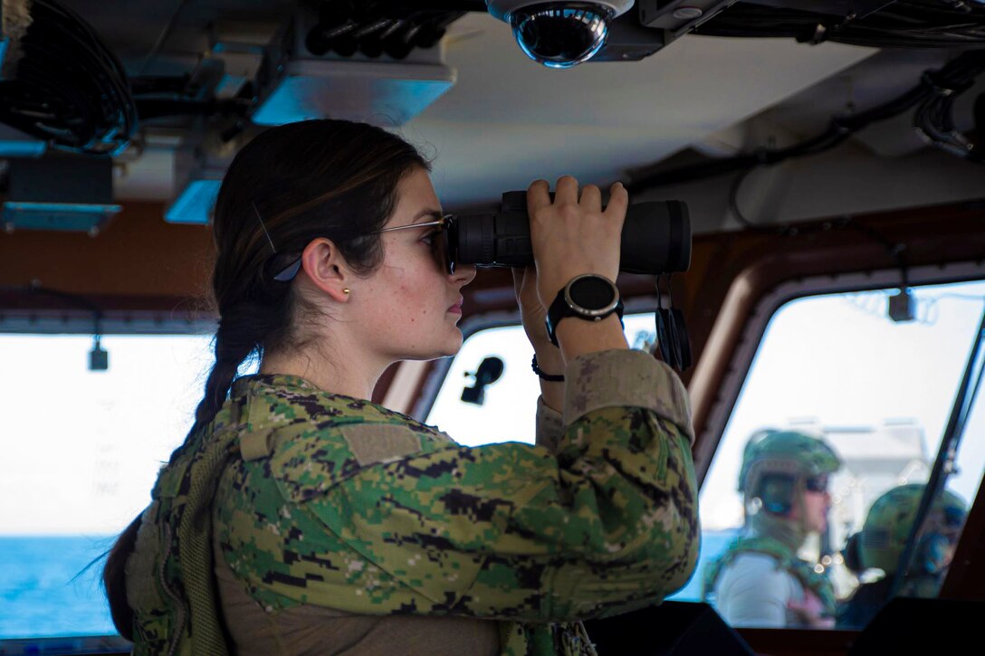 A coast guardsman looks through binoculars aboard a ship.