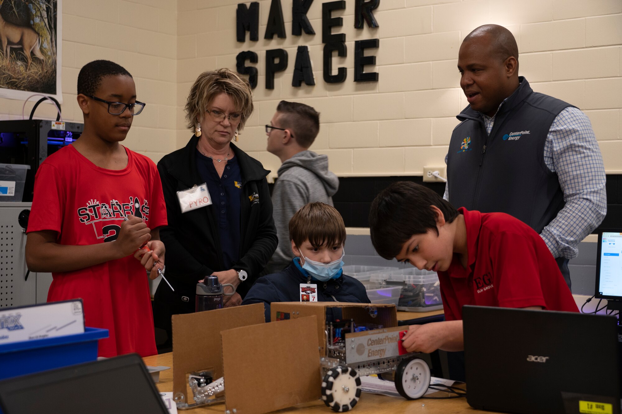 Two adults stand with three children working on a robot in a classroom.