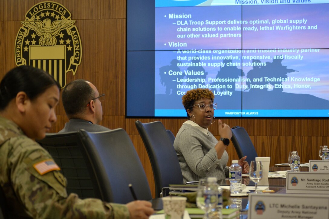 A woman speaks as two others listen. In the background, slides are being presented and a Defense Logistics Agency emblem adorns the wall.