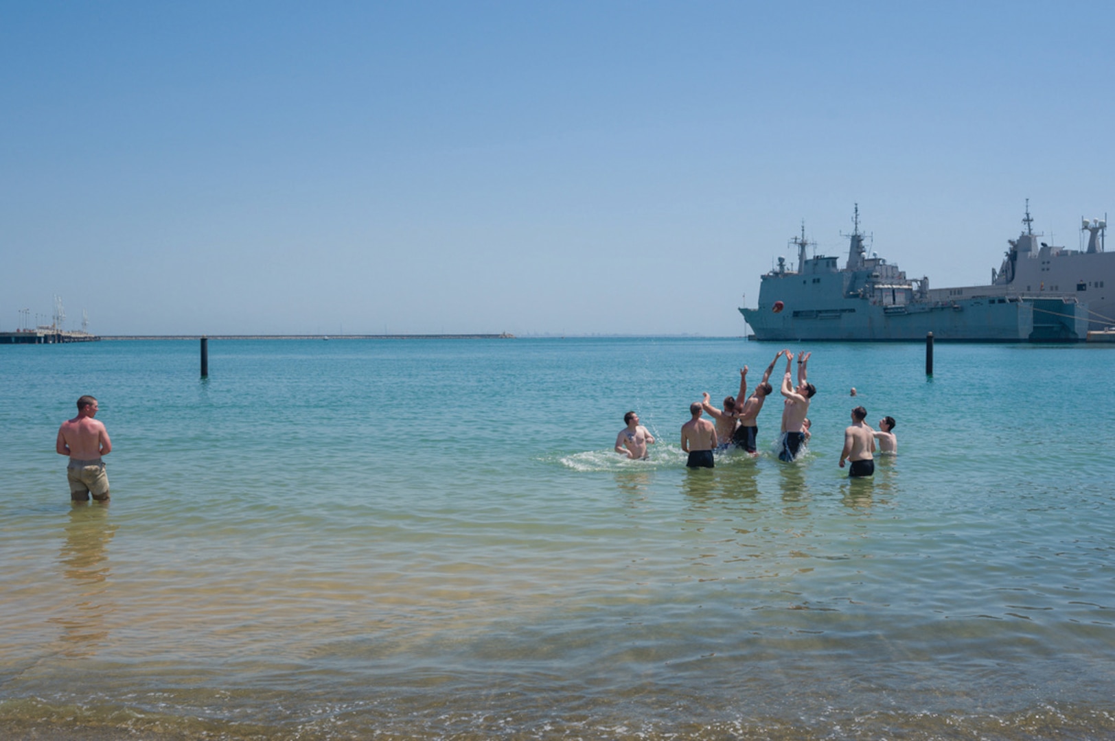 Men splash around on a beach with a Navy ship in the background.