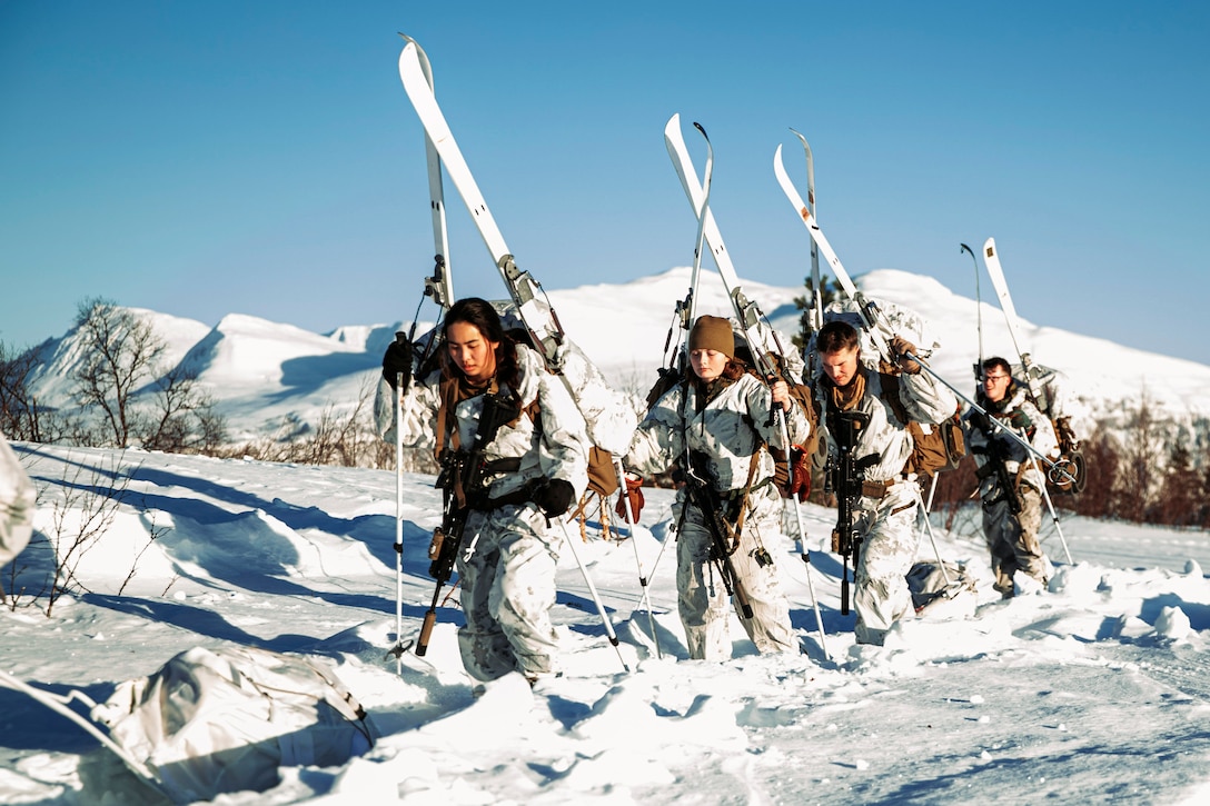 Four Marines hike up a snowy mountain while carrying skis.