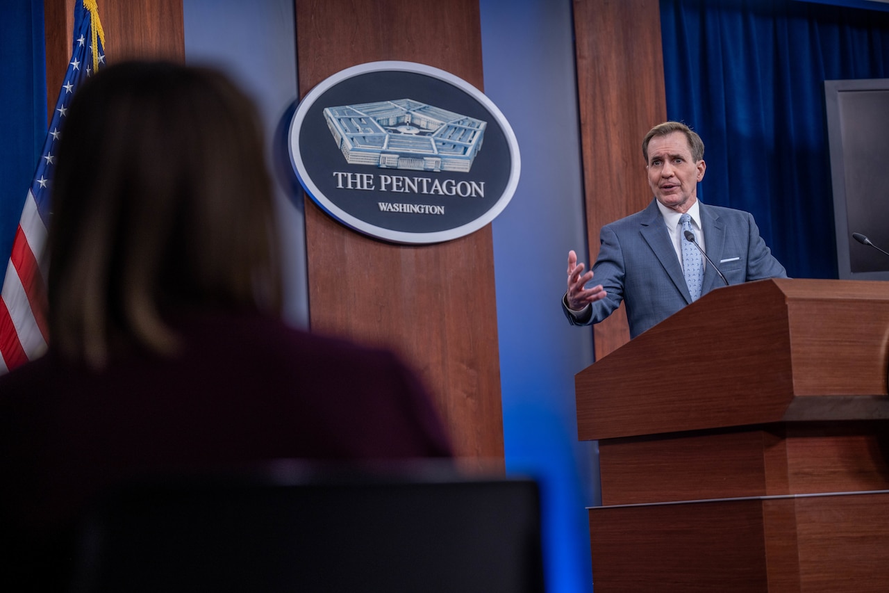 A man standing at a lectern speaks to members of the press.