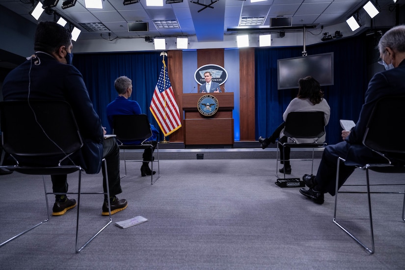 A man standing at a lectern speaks to members of the press.