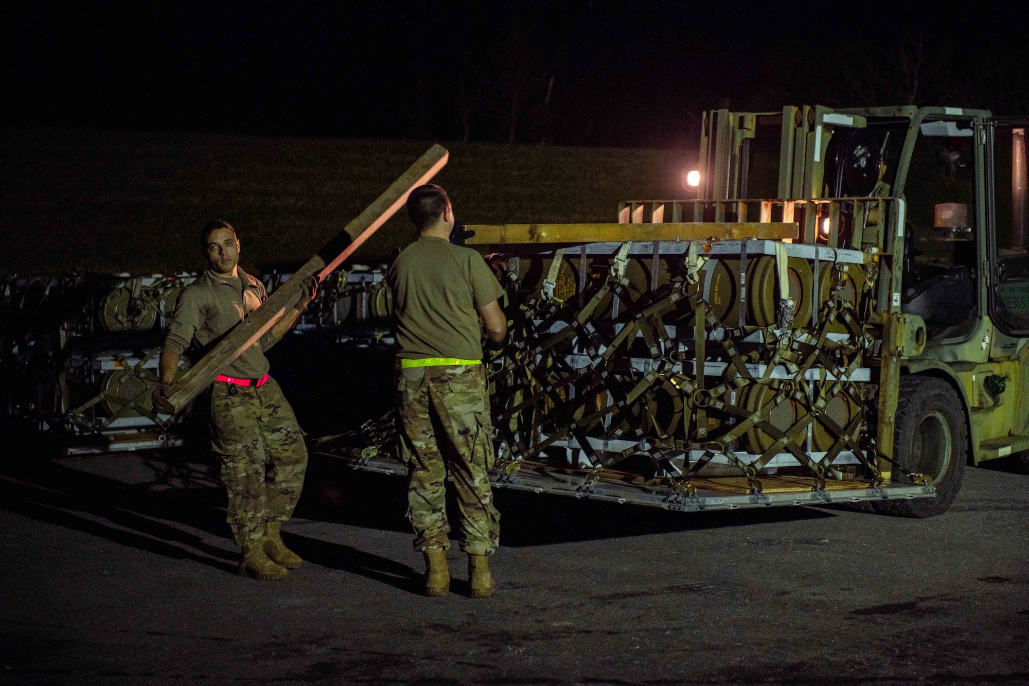 Two Airmen remove wooden posts from below a pallet and move them on top at night