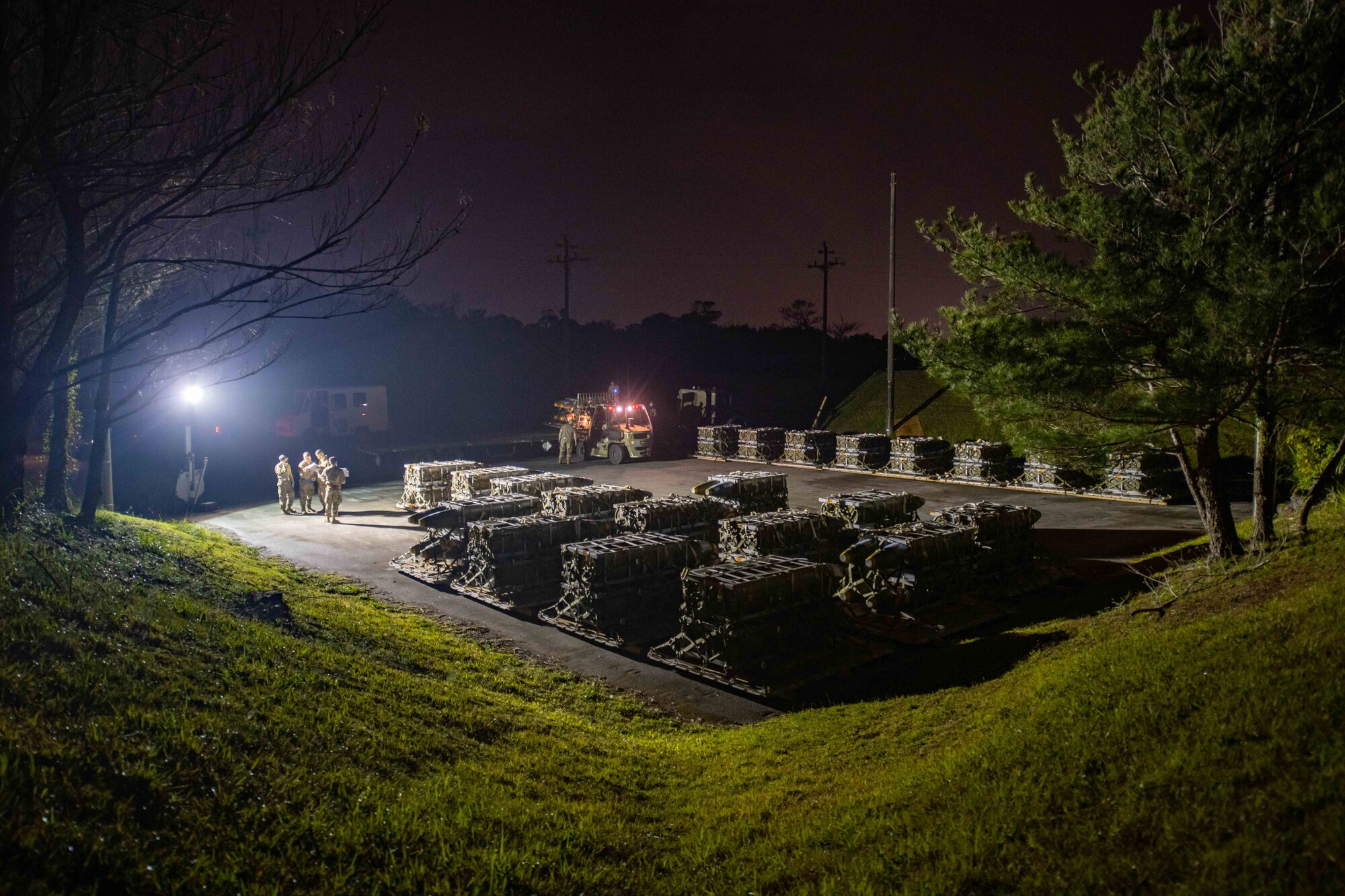 A pile of munitions sit in a parking lot at night, lit by lights and prepared on pallets ready to be loaded onto a truck