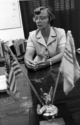 Black and white photo of woman in suit sitting at the table with flags setting