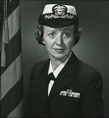 Black and white official portrait photo of woman in navy uniform with USA flag in background