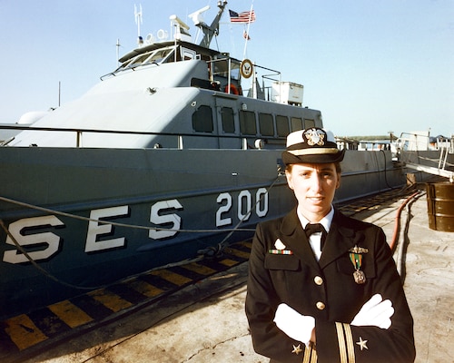 Woman in her uniform crossing arms, smiling at camera with USS Mount Vernon behind her