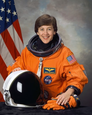 Official portrait of woman in her astronaut uniform smiling at camera with USA flag in background