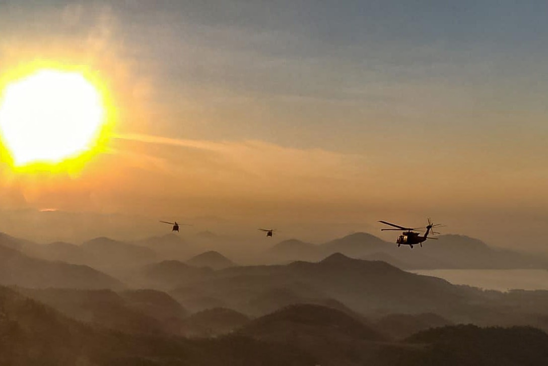 Three helicopters fly over hazy mountains toward the sun.