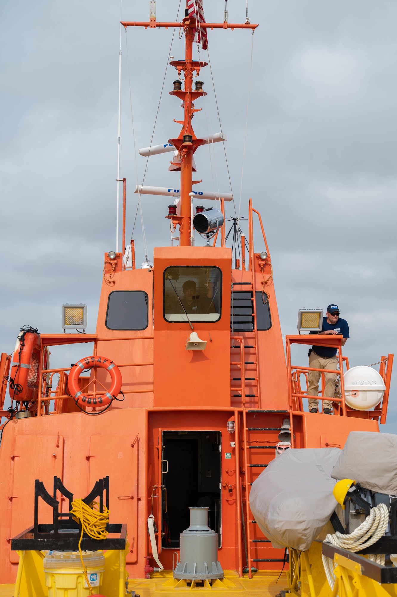 Members of a missile retriever boat crew man the vessel, with one standing on the dock while the other can be seen in the captains seat.