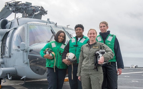 Women in their bright green jacket along with one woman in olive green uniform, they all holding their helmets smiling at camera with helicopter in background