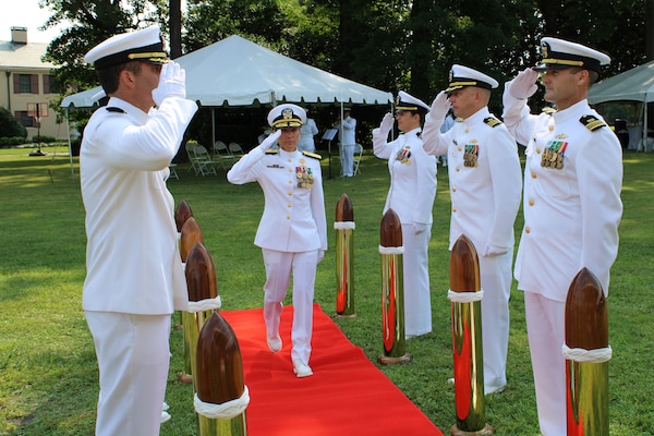 Woman in white navy uniform saluting as she walk through 2 rows of commanders in white uniform saluting