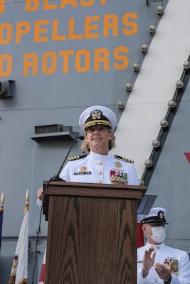 Woman in her white navy uniform giving speech at pedestal with ship behind her