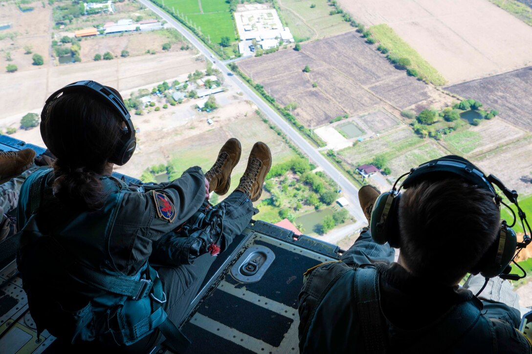 Two airmen sit inside an airborne aircraft looking over land.